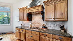 Kitchen with light wood-type flooring, baseboards, stainless steel gas stovetop, backsplash, and dark stone countertops