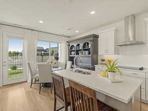 Kitchen with a sink, wall chimney range hood, a kitchen island with sink, black electric cooktop, and light wood-style flooring