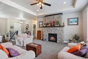 Carpeted living area featuring a raised ceiling, baseboards, ceiling fan, a fireplace, and a chandelier