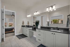 Full bath with marble look tiles, two vanities, ceiling fan, ensuite bath, and baseboards