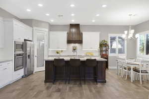 Kitchen featuring stainless steel appliances, custom range hood, a chandelier, an island with sink, and light wood-type flooring