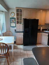 Kitchen featuring black fridge with ice dispenser, a sink, light tile patterned floors, dark countertops, and glass insert cabinets