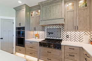 Kitchen featuring stainless steel gas cooktop, multiple ovens, backsplash, glass insert cabinets, and light brown cabinetry