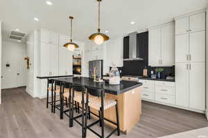 Kitchen with wall chimney exhaust hood, dark countertops, light wood finished floors, a kitchen breakfast bar, and recessed lighting