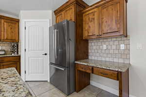 Kitchen featuring freestanding refrigerator, light tile patterned floors, tasteful backsplash, light stone counters, and brown cabinets