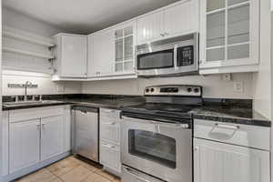Kitchen featuring appliances with granite countertops, stainless steel finishes, a sink, open shelves, light travertine tile floors, and white cabinetry