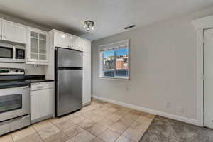 Kitchen with appliances with stainless steel finishes, glass insert cabinets, granite countertops, white cabinetry, and light travertine floors