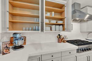 Kitchen featuring wall chimney exhaust hood, stainless steel gas stovetop, open shelves, tasteful backsplash, and light countertops