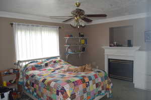 Carpeted bedroom with crown molding, a textured ceiling, a glass covered fireplace, and ceiling fan