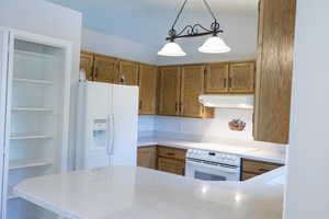 Kitchen with under cabinet range hood, white appliances, brown cabinetry, hanging light fixtures, and light countertops