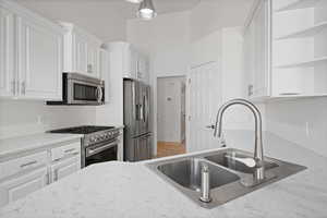 Kitchen with stainless steel appliances, a sink, open shelves, white cabinets, and light stone counters
