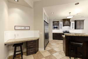 Kitchen with built in appliances, a breakfast bar area, dark brown cabinets, custom range hood, and baseboards