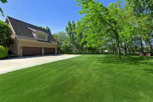 View of green lawn with a garage, concrete driveway, and view of wooded area