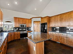 Kitchen with appliances with stainless steel finishes, a kitchen island, decorative backsplash, stone countertops, and vaulted ceiling