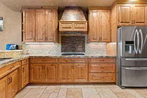 Kitchen featuring appliances with stainless steel finishes, custom range hood, tasteful backsplash, stone tile floors, and light stone countertops