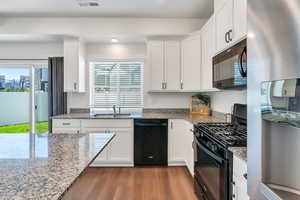 Kitchen with black appliances, wood finished floors, white cabinets, a sink, and light stone counters
