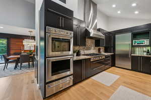 Kitchen featuring built in appliances, a warming drawer, dark cabinets, wall chimney exhaust hood, and light wood-type flooring