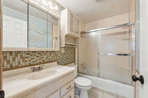 Full bath with a textured ceiling, enclosed tub / shower combo, vanity, toilet, and backsplash