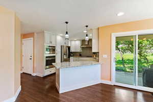 Kitchen with stainless steel appliances, wall chimney exhaust hood, a peninsula, light stone counters, and dark wood-type flooring