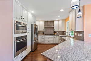 Kitchen with stainless steel appliances, wall chimney range hood, light stone counters, dark wood-style floors, and decorative backsplash