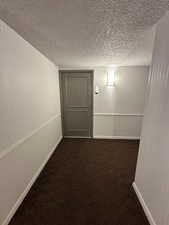 Hallway with a textured ceiling, dark colored carpet, and baseboards