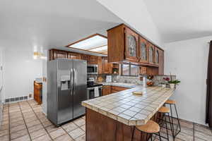 Kitchen with a peninsula, tile countertops, decorative backsplash, brown cabinetry, and appliances with stainless steel finishes