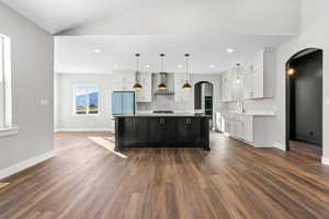 Kitchen featuring wall chimney exhaust hood, arched walkways, white cabinetry, recessed lighting, and vaulted ceiling
