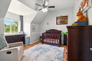 Bedroom featuring wood finished floors, a nursery area, lofted ceiling, a ceiling fan, and baseboards
