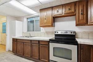 Basement secondary kitchen with stainless steel range with electric cooktop, a sink, tile countertops, light wood-style floors, and brown cabinets