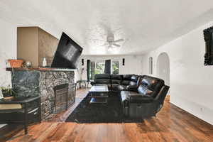 Living area with arched walkways, ceiling fan, wood-type flooring, a textured ceiling, and a stone fireplace
