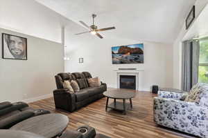 Living room featuring vaulted ceiling, a glass covered fireplace, a ceiling fan, wood finished floors, and baseboards
