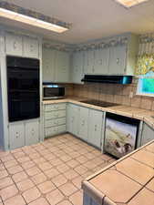 Kitchen with black appliances, under cabinet range hood, decorative backsplash, and tile counters