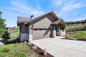 View of front of property featuring stone siding, concrete driveway, an attached garage, a chimney, and roof with shingles