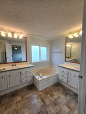 Full bath with two vanities, stone finish floors, a garden tub, crown molding, and a textured ceiling
