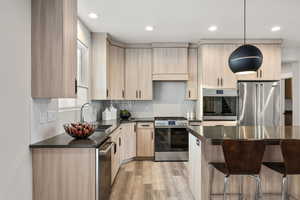 Kitchen with light brown cabinetry, backsplash, appliances with stainless steel finishes, hanging light fixtures, and light wood-style floors