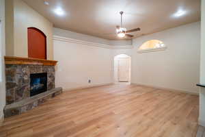 Unfurnished living room featuring a ceiling fan, a stone fireplace, light wood-type flooring, arched walkways, and baseboards