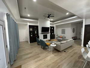 Living room featuring light wood-type flooring, ceiling fan, a tray ceiling, baseboards, and a fireplace