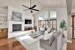 Living room with a glass covered fireplace, a ceiling fan, light wood finished floors, built in shelves, and high vaulted ceiling