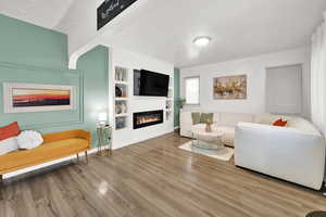 Living area featuring a glass covered fireplace, wood finished floors, a textured ceiling, built in features, and baseboards