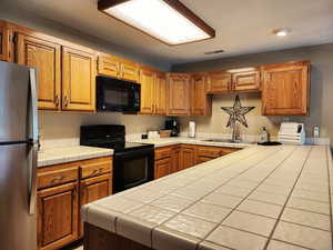 Kitchen with black appliances, tile countertops, and brown cabinetry
