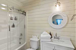 Bathroom with wood walls, vanity, shower / bath combination with glass door, and toilet