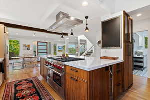 Kitchen with premium appliances, wall chimney range hood, light wood-type flooring, light countertops, and decorative light fixtures
