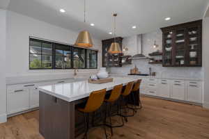 Kitchen featuring wall chimney range hood, light countertops, backsplash, a kitchen island, and glass insert cabinets