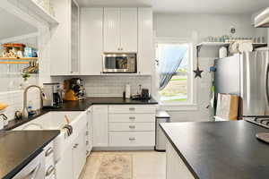 Kitchen with appliances with stainless steel finishes, dark countertops, tasteful backsplash, white cabinetry, and glass insert cabinets