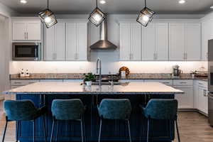 Kitchen featuring stainless steel microwave, wall chimney range hood, wood finished floors, an island with sink, and white cabinets