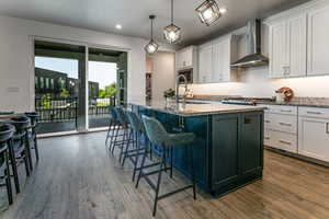 Kitchen featuring wall chimney exhaust hood, an island with sink, dark wood-style flooring, white cabinets, and stone countertops