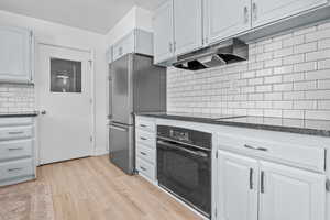 Kitchen featuring black appliances, under cabinet range hood, light wood-style flooring, tasteful backsplash, and white cabinetry