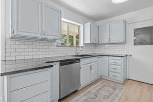 Kitchen with stainless steel dishwasher, a sink, light wood-type flooring, backsplash, and dark stone countertops