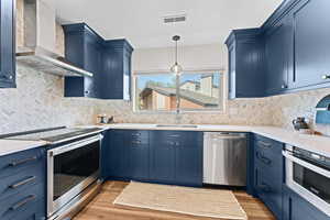 Kitchen with appliances with stainless steel finishes, wall chimney exhaust hood, light wood finished floors, and blue cabinetry