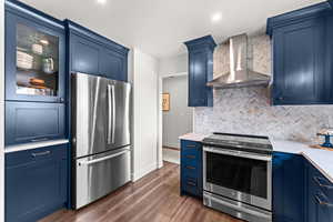 Kitchen with stainless steel appliances, wall chimney exhaust hood, blue cabinets, light countertops, and recessed lighting